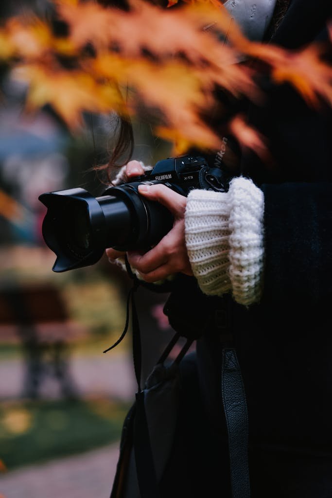 A young woman captures a moment outdoors with a camera amidst autumn foliage.